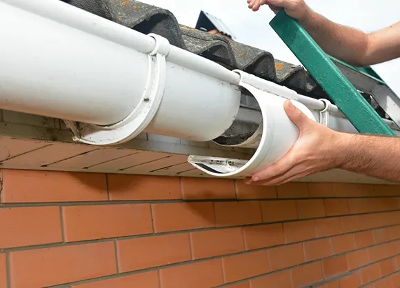 Person cleaning white gutters attached to a brick house using a pressure washer in Brockton, MA